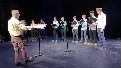 Neuf étudiant·es chantant sur scène avec un chef d'orchestre qui les guide.