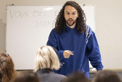 Un jeune homme en train de parler dans une salle de classe, devant un tableau.