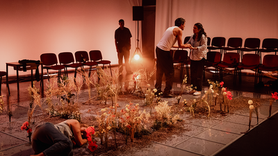 Une femme est couchée dans un parterre de fleurs au premier plan. Un couple semble se disputer à l'arrière plan.