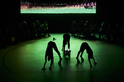 Trois danseuses vêtues de pieds de boucs au centre d'une scène, entourent un chien-robot.